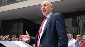 Man in suit and red tie speaking at a podium to an audience in a modern building.