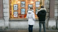 Two people looking at real estate listings in an estate agents window, showcasing properties for sale.