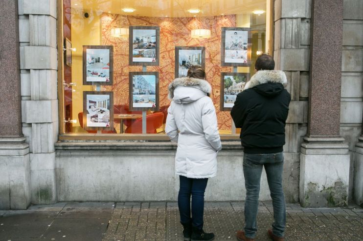 Two people looking at real estate listings in an estate agents window, showcasing properties for sale.