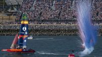 SailGP F50 catamaran Los Gallos sailing past a cheering crowd as red and blue smoke erupts.