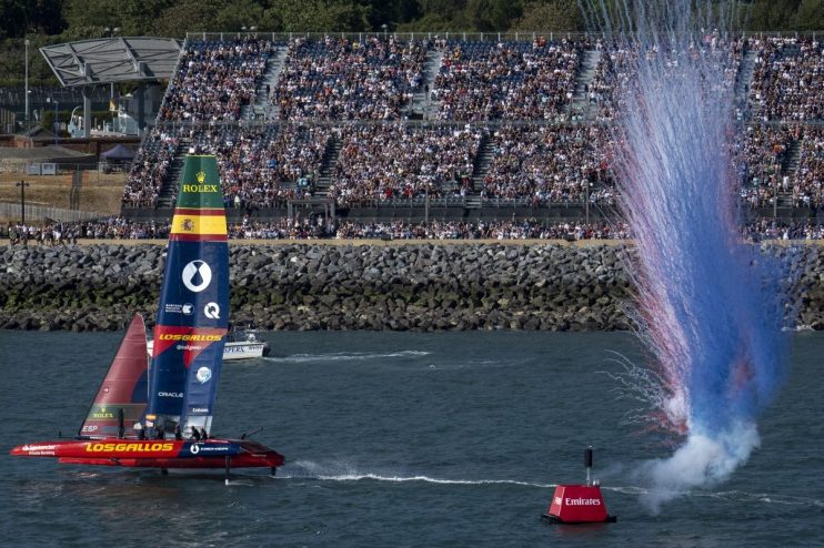 SailGP F50 catamaran Los Gallos sailing past a cheering crowd as red and blue smoke erupts.