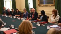 John Healey, an MP, speaks at a formal business meeting with other politicians and executives around a green table.