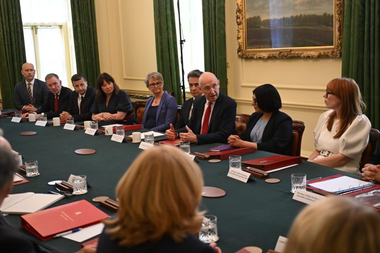 John Healey, an MP, speaks at a formal business meeting with other politicians and executives around a green table.