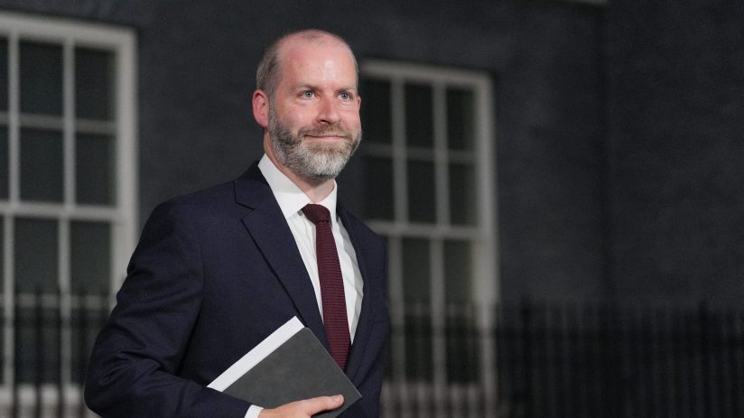 Jonathan Reynolds, Labour MP, in a suit and tie, holding a notebook, looking right, with a building behind him