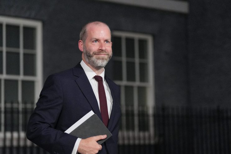 Jonathan Reynolds, Labour MP, in a suit and tie, holding a notebook, looking right, with a building behind him