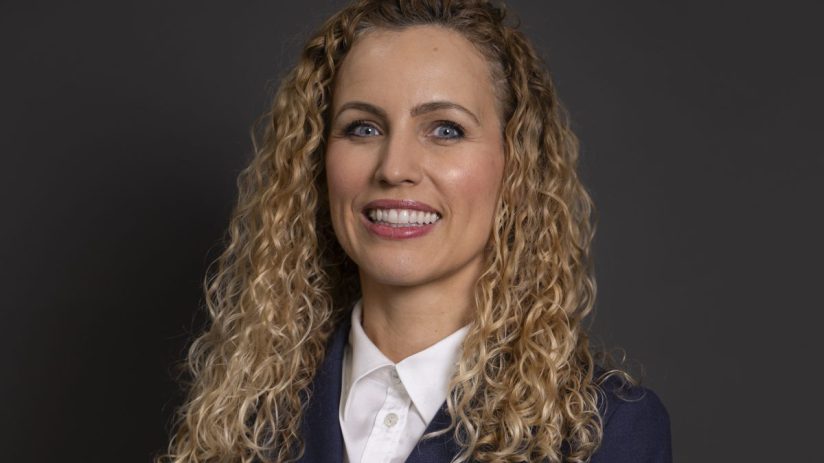 Katie White, a woman with curly blonde hair, smiling in a navy blazer and white collared shirt against a dark background.