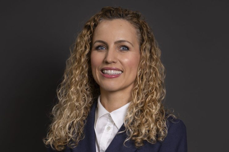 Katie White, a woman with curly blonde hair, smiling in a navy blazer and white collared shirt against a dark background.