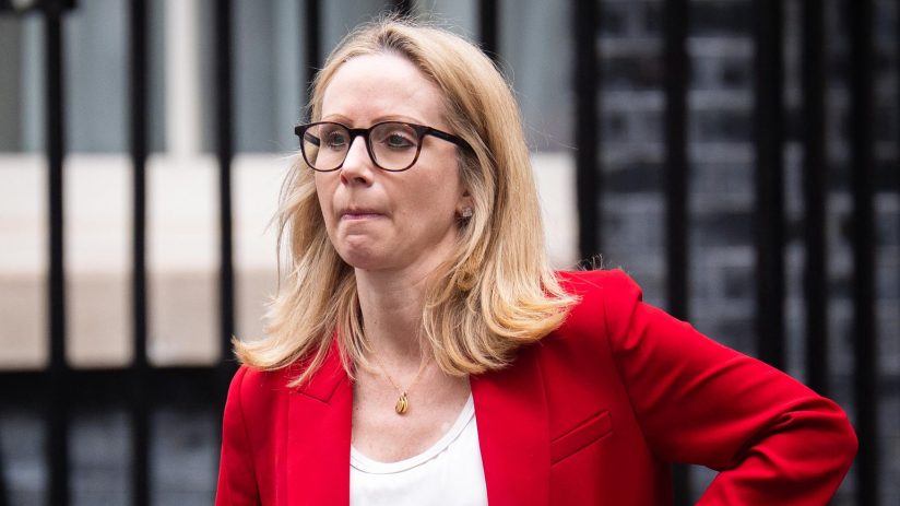 Lucy Rigby, a blonde woman in glasses and a red blazer, with a phone in her pocket, walking past a black gate.