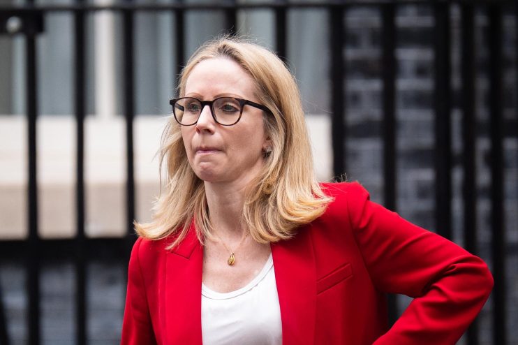 Lucy Rigby, a blonde woman in glasses and a red blazer, with a phone in her pocket, walking past a black gate.