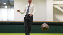Bald man in glasses and a tie kicking a soccer ball on an indoor artificial turf field.