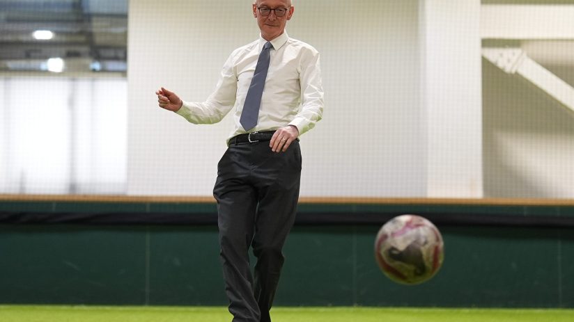 Bald man in glasses and a tie kicking a soccer ball on an indoor artificial turf field.