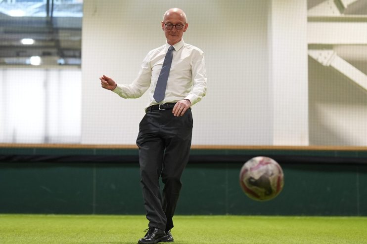 Bald man in glasses and a tie kicking a soccer ball on an indoor artificial turf field.
