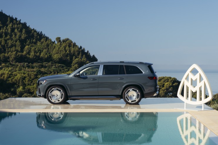 Gray Mercedes-Maybach GLS SUV parked next to a pool with a Maybach logo, green hills, and ocean in background