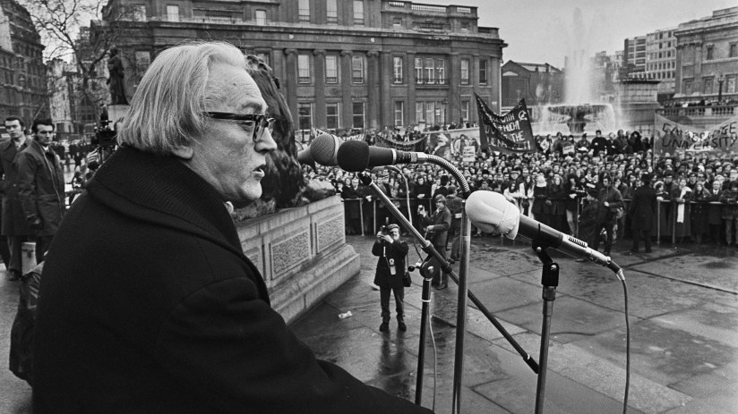 Michael Foot speaking at a public rally in a city square with a large crowd and buildings in the background.