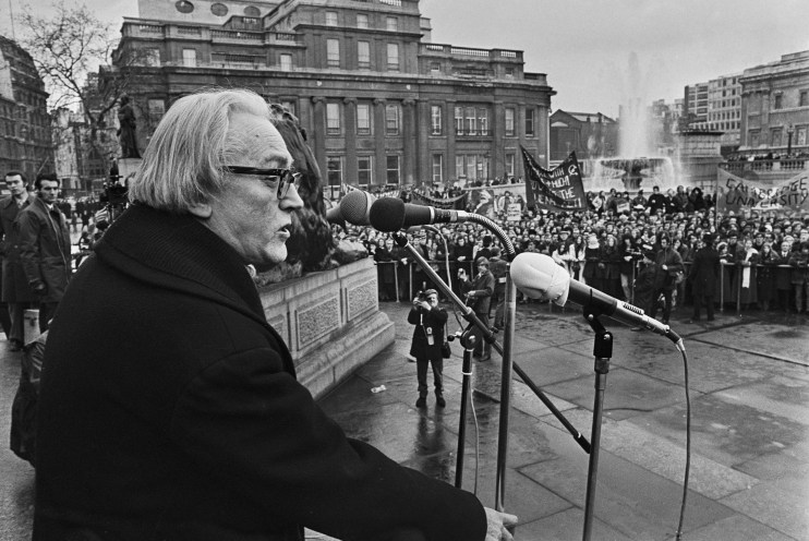 Michael Foot speaking at a public rally in a city square with a large crowd and buildings in the background.