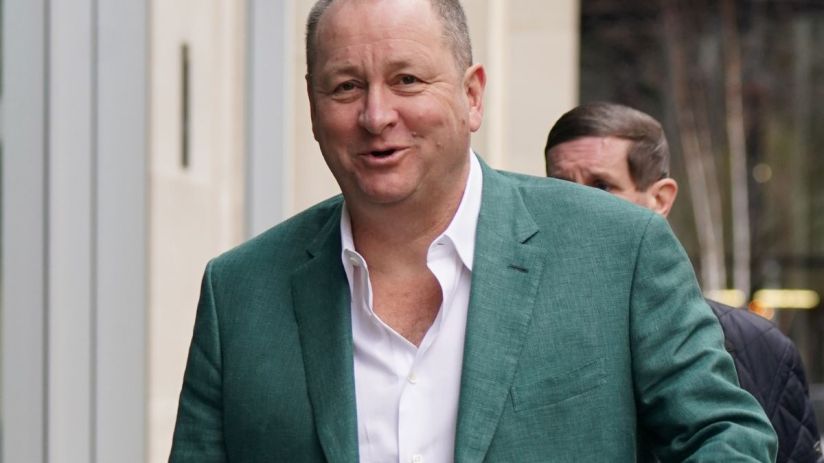 Mike Ashley in a business suit at a corporate event, discussing strategic plans, surrounded by executives and media personnel