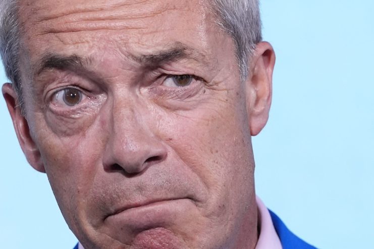 Nigel Farage speaking at a podium, dressed in a suit, addressing an audience at a business conference event