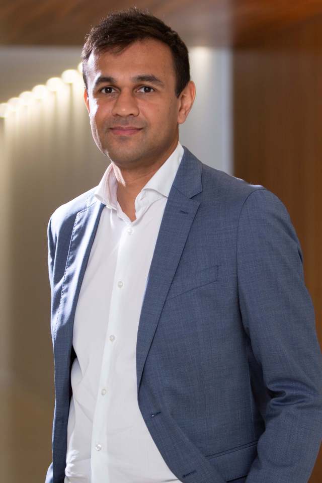 Nimesh, a professional man in a blue suit jacket and white shirt, smiling in a corporate setting.