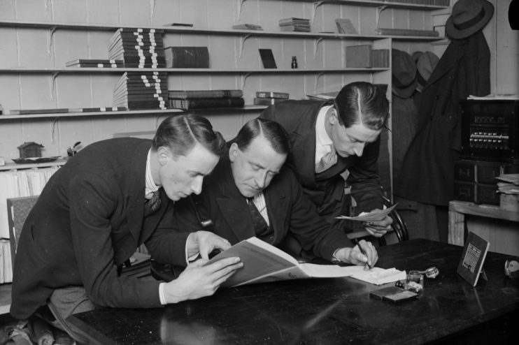 Diverse group of office workers collaborating at desks with laptops and paperwork in a modern, well-lit workspace.
