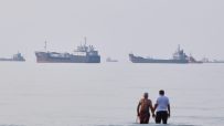 Two men stand in the ocean with multiple oil tankers and cargo ships in the hazy distance.