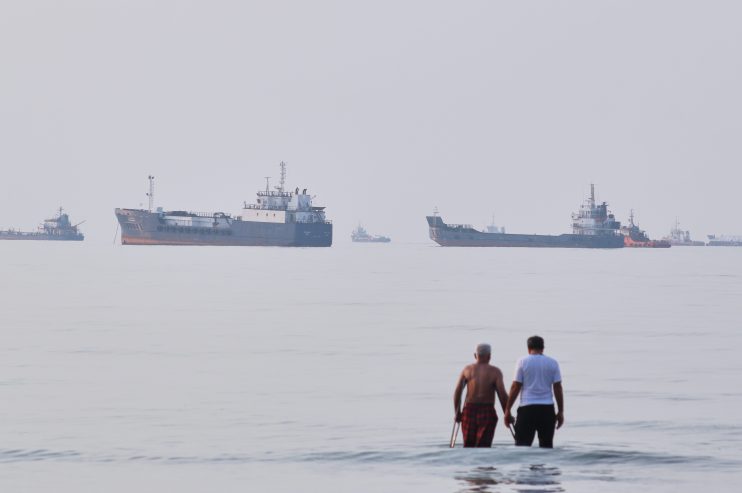 Two men stand in the ocean with multiple oil tankers and cargo ships in the hazy distance.