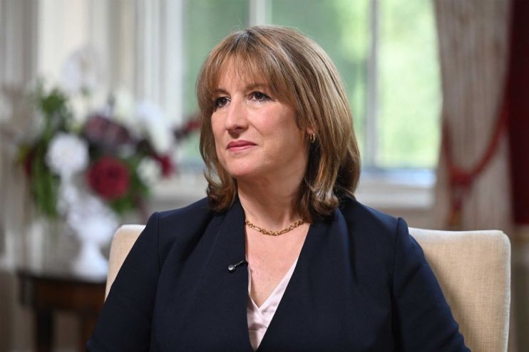 Rachel Reeves delivering a speech at a press event, wearing a navy blazer and standing in front of a backdrop with logos.