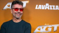 Simon Cowell smiling brightly during a press event, dressed in a classic tailored suit, showcasing his signature confident...