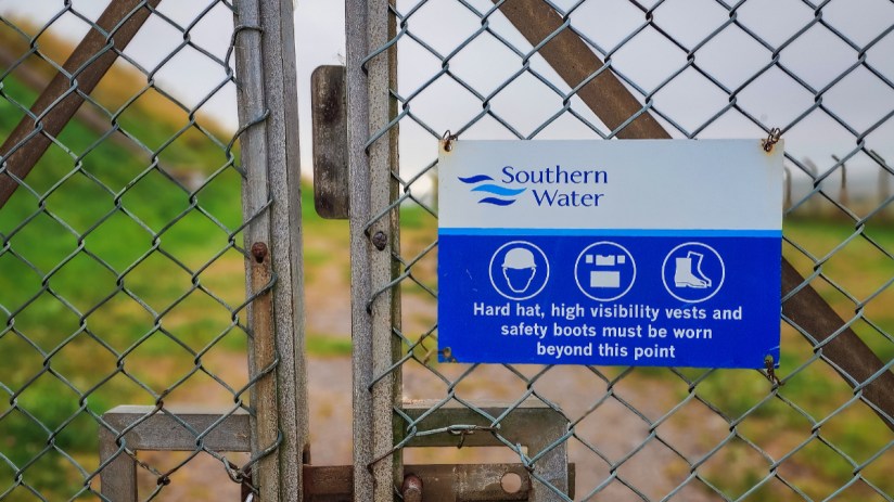Southern Water safety sign on a chain link fence, detailing required PPE like hard hats and safety boots.