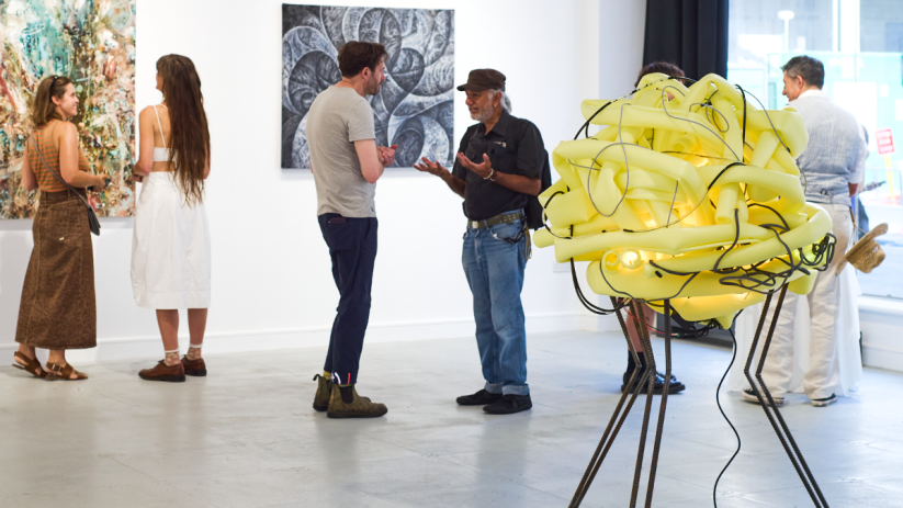 Art enthusiasts view colorful abstract paintings and a unique yellow light sculpture at a Square Mile art gallery.