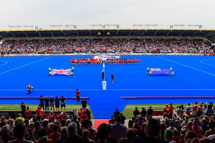 Teams and flags on a blue hockey pitch before a match, with a stadium full of spectators.