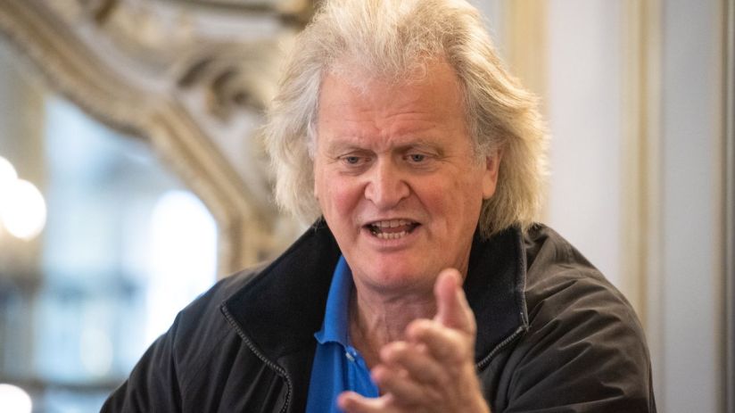 Tim Martin, founder of JD Wetherspoon, speaking and gesturing with an open hand, wearing a blue polo shirt and dark jacket.