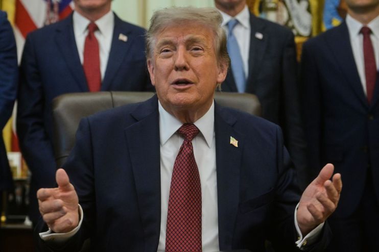 Donald Trump speaking at a desk, gesturing with hands, wearing a dark suit and red tie.