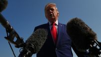 Donald Trump speaking at a press conference with microphones, blue sky background