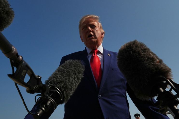 Donald Trump speaking at a press conference with microphones, blue sky background