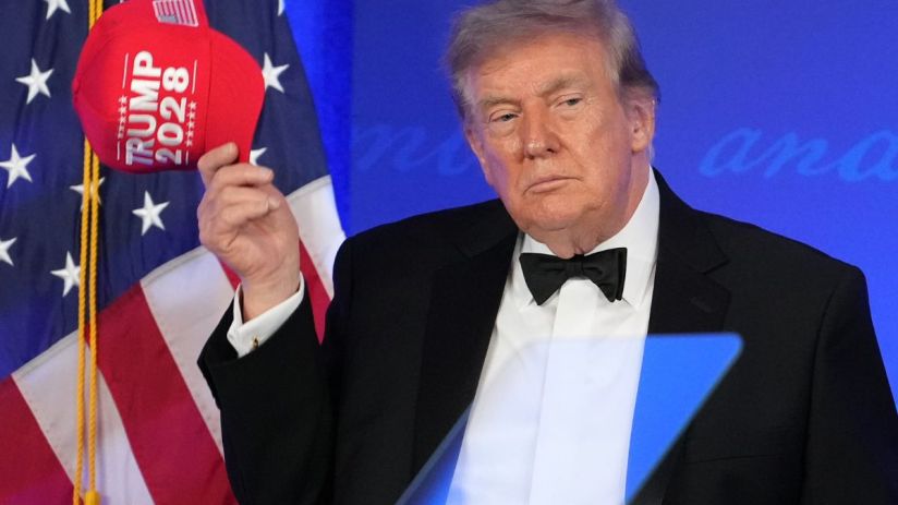 Donald Trump holding a red TRUMP 2028 hat, wearing a tuxedo with an American flag in the background