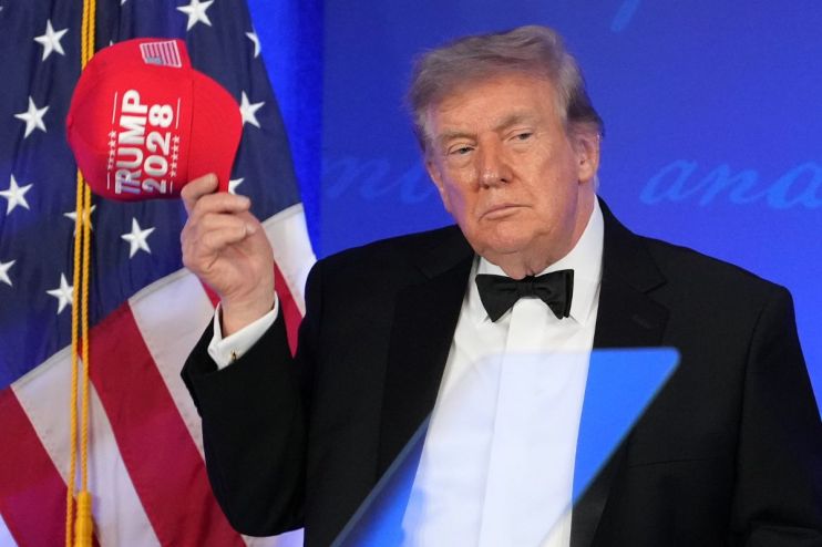 Donald Trump holding a red TRUMP 2028 hat, wearing a tuxedo with an American flag in the background