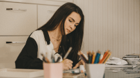 Young woman drawing or writing at a desk, surrounded by art supplies, illustrating human creativity vs AI.