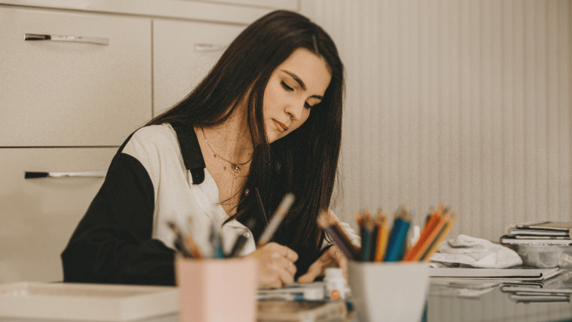 Young woman drawing or writing at a desk, surrounded by art supplies, illustrating human creativity vs AI.