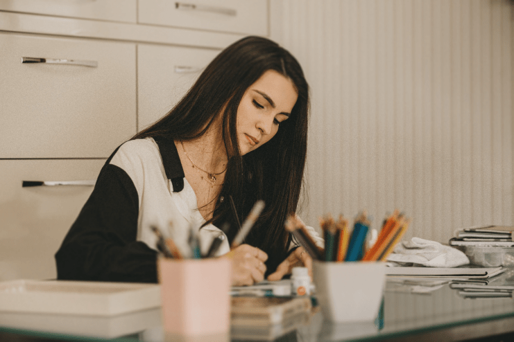 Young woman drawing or writing at a desk, surrounded by art supplies, illustrating human creativity vs AI.