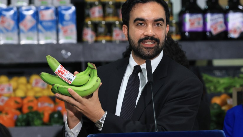 Zohran Mamdani, a man with a beard, holds a bunch of green bananas with a 30% off label at a grocery store.