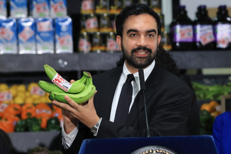 Zohran Mamdani, a man with a beard, holds a bunch of green bananas with a 30% off label at a grocery store.