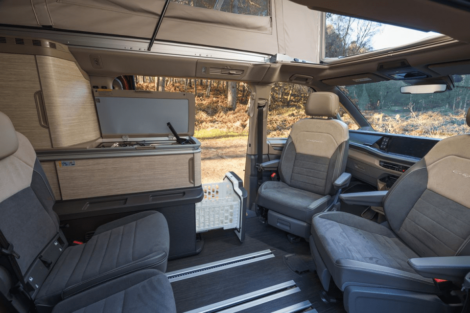Interior of a modern camper van with swivel front seats, pop-up roof, and a compact kitchen unit with an open lid.