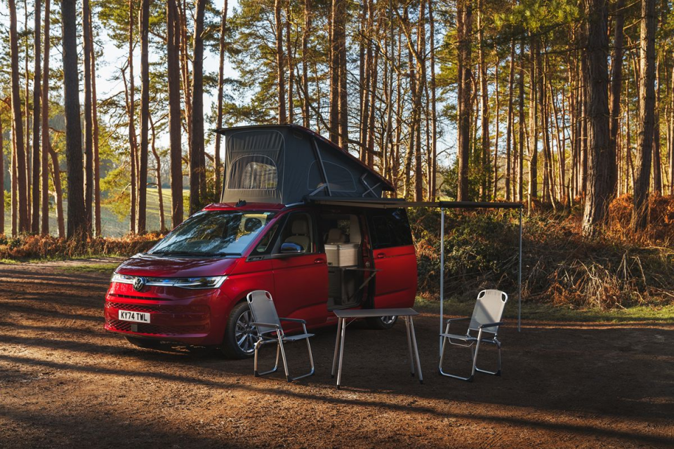 Red VW California camper van with pop-top roof and awning set up in a pine forest, with chairs and table.