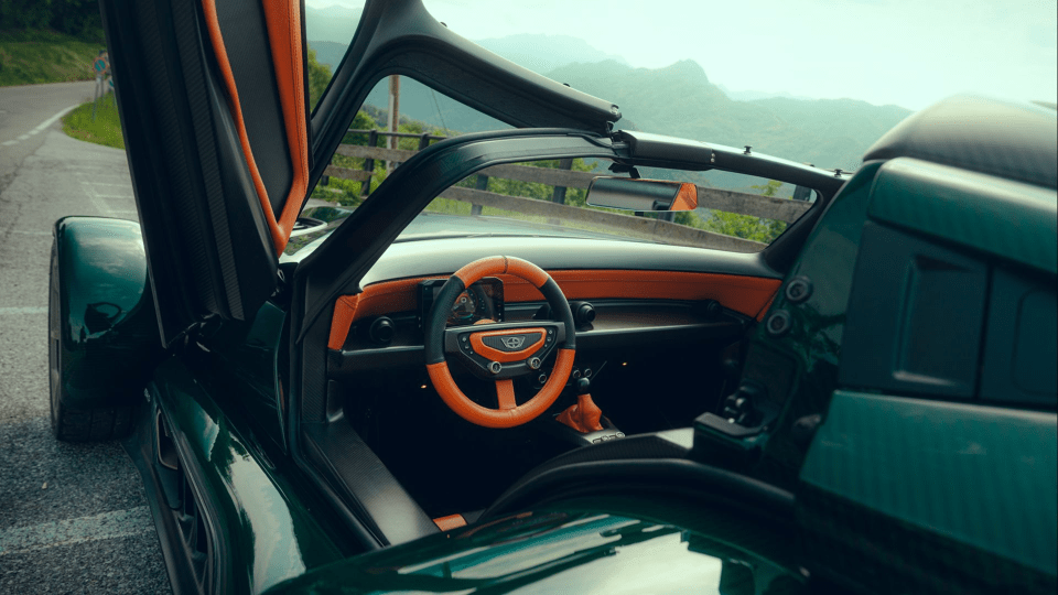 Interior of a green sports car with orange and black steering wheel, open door, mountain road background
