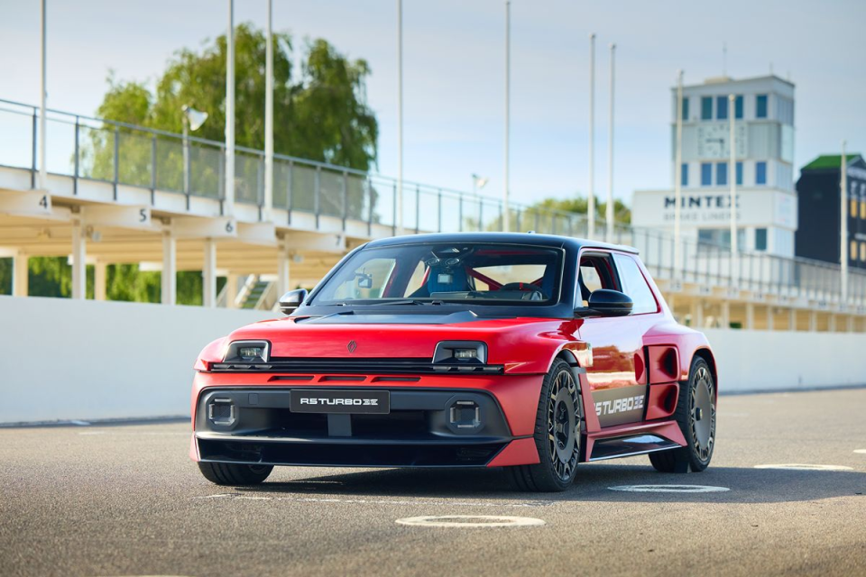 Red and black Renault R5 Turbo 3E concept car on a racetrack, with grandstands and a control tower in background.