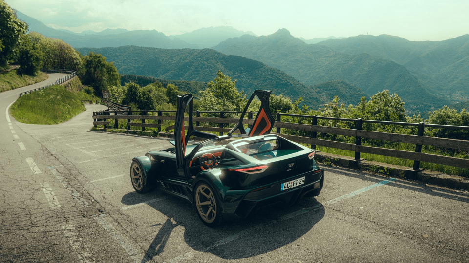 Dark green sports car with open doors parked on a mountain road overlooking a scenic valley.