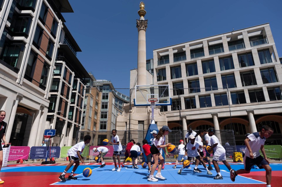Children playing basketball at a pop-up court during the London Sports Festival, with buildings and a monument in the back...