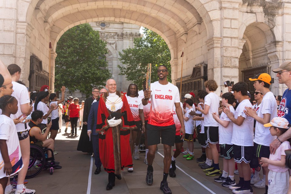 A man in a red and black ceremonial robe walks with a man holding the Kings Baton through a stone archway, surrounded by c...
