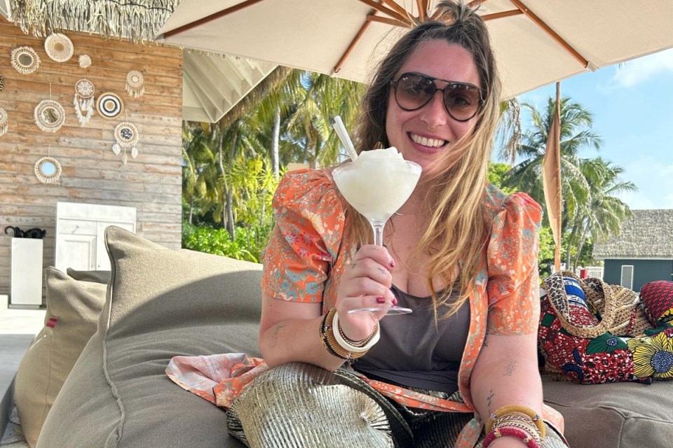 Woman in sunglasses smiles, holding a frozen drink at a Maldives Ifuru bar with palm trees and decor.