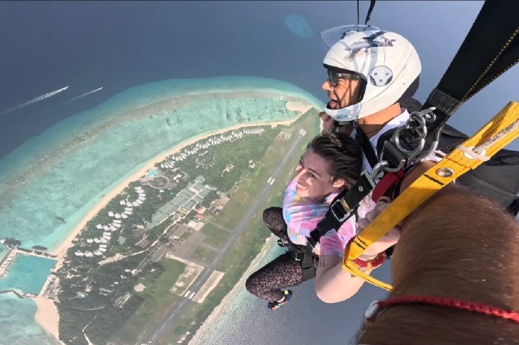 Two people skydiving over the Maldives, with turquoise water, coral reefs, and an island resort below.